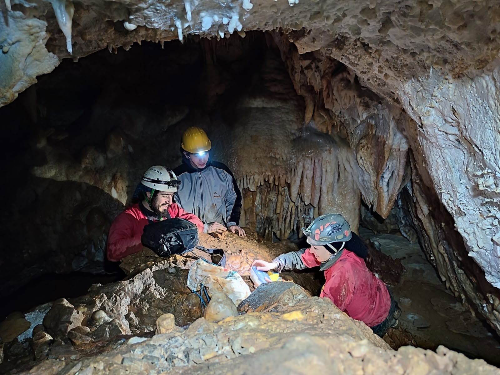Tre ricercatori con casco e frontale raccolgono campioni tra stalattiti e concrezioni in una grotta